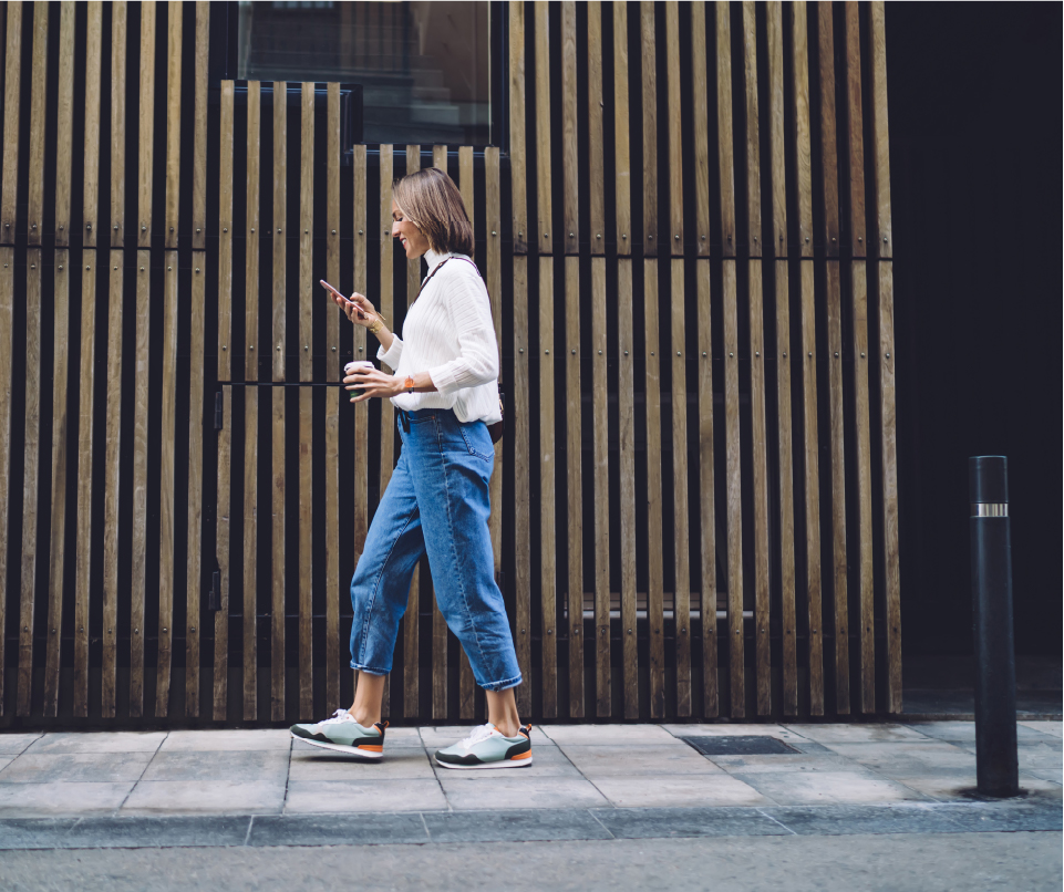Woman carrying coffee while waking in the street and smiling while looking at her mobile device